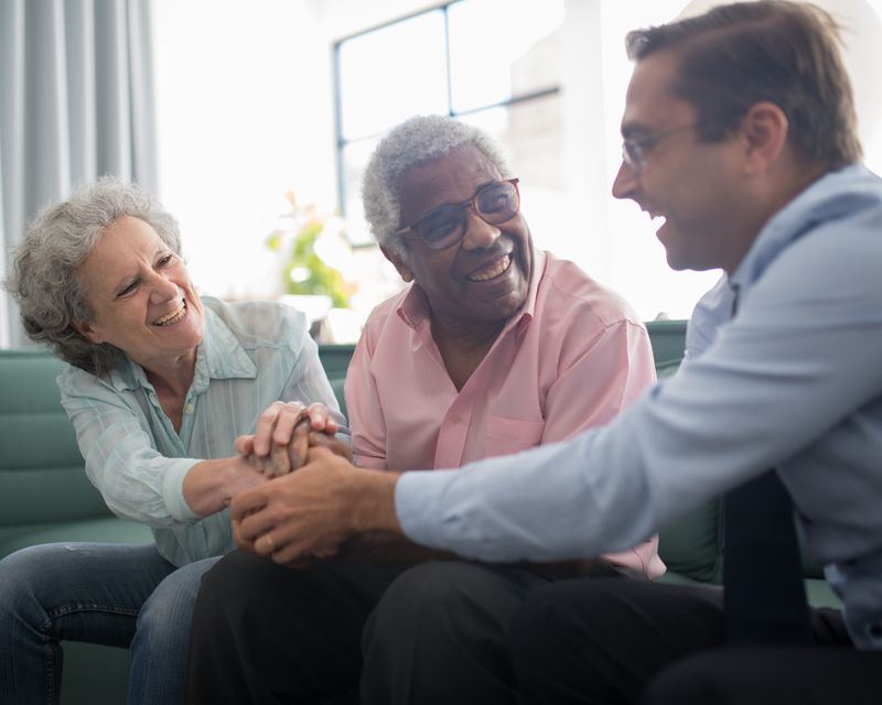 An older woman and an older man laughing and holding hands with a visitor at home